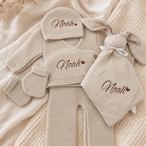 Op de afbeelding: Een beige gebreide babyset bestaande uit een overslagtop met lange mouwen, een broekje, een mutsje en slofjes, allemaal geborduurd met de naam Noah en een klein hartje. Een bijpassend beige mousseline knuffeldoekje met konijnenkop is inbegrepen.