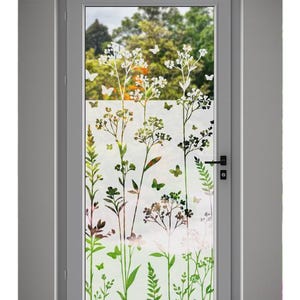 Può includere: Una porta con cornice bianca con un pannello di vetro smerigliato con un design botanico. Il design include fiori bianchi, foglie verdi e farfalle. Lo sfondo mostra una vista sfocata di alberi e fogliame.
