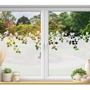 Könnte beinhalten: Ein weißes Fenster mit einem dekorativen, mattierten Blattdesign. Das Fenster bietet einen Blick auf eine grüne Landschaft. Ein Holzregal beherbergt eine Vase mit getrockneten Pflanzen, eine Sukkulente, eine Kerze und ein Tablett mit einer Flasche, Pflanzen und einer Kerze.