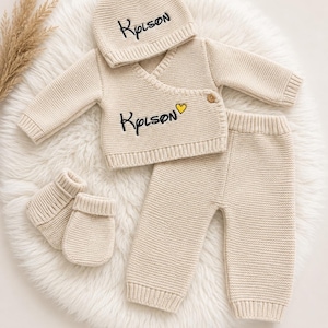 Peut inclure: Un ensemble tricoté beige pour bébé comprenant un gilet cache-cœur à manches longues, un pantalon, un bonnet et des moufles. Le gilet et le bonnet portent le nom Kpison brodé en noir, avec un petit cœur jaune à côté du nom sur le gilet.