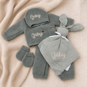 Può includere: Un set in maglia color verde salvia per neonati che include un maglione a maniche lunghe, pantaloni, un berretto e muffole, tutti con il nome Gaby ricamato. È incluso un doudou in mussola verde salvia con testa di coniglio e il nome Gaby.