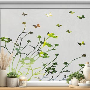 May include: A window with a floral and butterfly design. The design features green and yellow flowers and butterflies against a frosted background. Decorative items, including a vase with dried plants, a succulent, and candles, are on the windowsill.