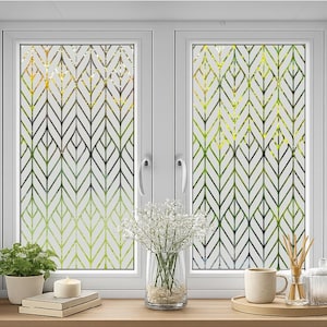 Può includere: Una finestra bianca con un motivo decorativo a foglie nei toni del verde, giallo e nero. La finestra è incorniciata in bianco e ha una mensola in legno con una piccola pianta in vaso, una candela, libri e una tazza di caffè.