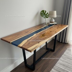Puede incluir: Una mesa rectangular de madera con un río de resina azul oscuro en el centro. La mesa tiene un acabado de vetas de madera natural y patas de metal negro. Una lámpara, libros y una planta están sobre la mesa.