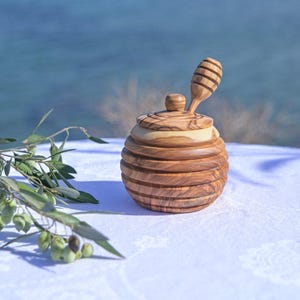 Könnte beinhalten: Ein hölzerner Honigtopf mit passendem Dipper, aus Olivenholz gefertigt. Der Topf hat ein rundes, geripptes Design und einen Deckel mit kleinem Knopf. Ein Olivenzweig liegt auf der weißen Tischdecke. Der Hintergrund ist eine verschwommene Ansicht des Meeres.