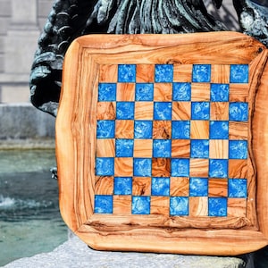 May include: A handcrafted wooden chessboard with a unique design. The board features a checkerboard pattern of natural wood squares and vibrant blue resin squares. The board is framed with a thick, natural wood border.