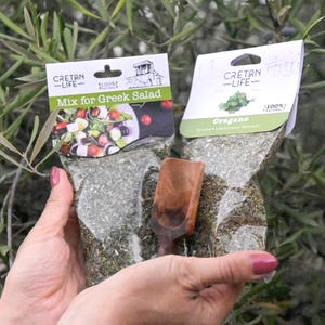 Peut inclure: Deux sachets transparents d'herbes Cretan Life, l'un étiqueté "Mix for Greek Salad" et l'autre "Oregano". Une petite cuillère en bois repose dans le sachet d'origan. Le fond est composé de feuillage vert.