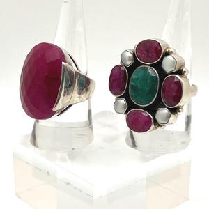 May include: Two silver rings displayed on clear stands. One ring features a large, faceted, oval-shaped ruby. The other ring has a cluster design with a central emerald surrounded by rubies and white stones, likely pearls.