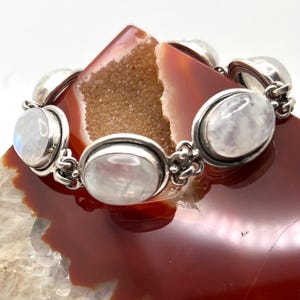 May include: A silver bracelet with oval moonstone cabochons. The bracelet features oval moonstones set in silver bezels, linked by decorative elements. The moonstones have a soft, milky iridescence. The bracelet is displayed on a red and brown stone.
