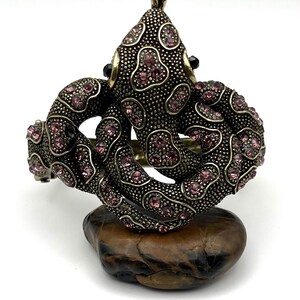 May include: An ornate, antique-style bracelet in a dark bronze tone, featuring a detailed design with pink rhinestones. The bracelet is shaped in a swirling pattern and rests on a small, brown stone. The overall aesthetic is vintage and glamorous.