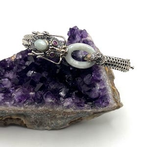 May include: A silver bracelet featuring a detailed dragon head with a pale green stone, small red accents, and clear pavé stones. The dragon holds a smooth, light green jade-like oval ring in its mouth, connected to a textured, scale-patterned band.