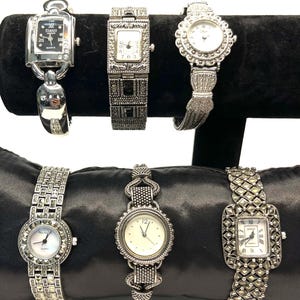 Pode incluir: Uma coleção de seis relógios prateados com vários designs. Os relógios apresentam mostradores quadrados, retangulares e redondos com algarismos romanos e movimento a quartzo. As pulseiras têm padrões e texturas intrincados, exibidos em uma superfície de veludo preto.