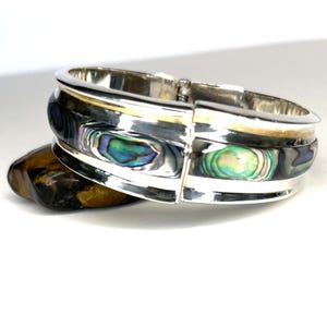 May include: A silver hinged bracelet with iridescent abalone shell inlays. The bracelet features a polished finish and a secure clasp. The abalone shell displays a range of colors, including blue, green, and purple. The bracelet is resting on a stone.