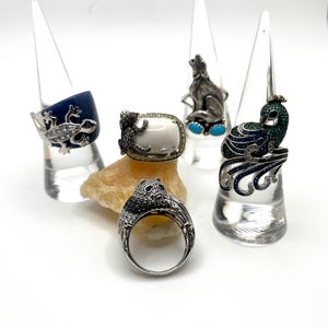 May include: An assortment of gemstone rings, each with a unique animal design. The rings feature a blue frog, a lizard on a white stone, a howling wolf, a peacock, and a panda bear. The rings are displayed on clear stands and a rock.