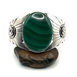 May include: A silver cuff bracelet with a large, oval malachite stone. The green stone is the focal point, set in a detailed silver band with floral designs on each side. The bracelet is displayed on a dark, textured surface.