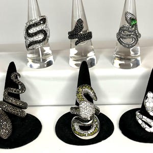 Op de afbeelding: Zes slangenringen worden getoond. De ringen hebben verschillende ontwerpen met zwarte, witte en groene edelstenen. Sommige ringen staan op transparante standaards, andere op zwarte fluwelen kegels. De ringen zijn een uniek sieraad.