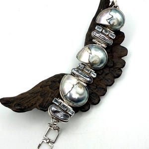 May include: A silver bracelet featuring three large, iridescent, semi-circular abalone shell segments alternating with smaller, elongated, dark grey shell pieces and small, light blue, rectangular faceted gemstones set in silver frames.