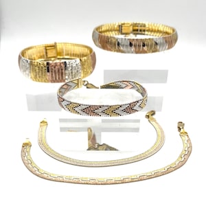 Puede incluir: Un surtido de pulseras y collares de oro, plata y oro rosa. Las pulseras presentan varios diseños, incluyendo una banda ancha y un patrón tejido. Los collares son de estilo cadena plana. Las joyas se exhiben sobre un fondo blanco.