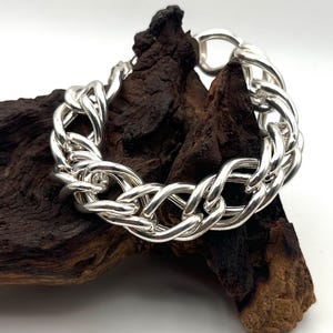 Puede incluir: Una pulsera de cadena de plata pulida con eslabones ovalados gruesos entrelazados que crean un patrón trenzado continuo. La pulsera tiene un acabado metálico altamente reflectante y un mecanismo de cierre circular en la parte superior.