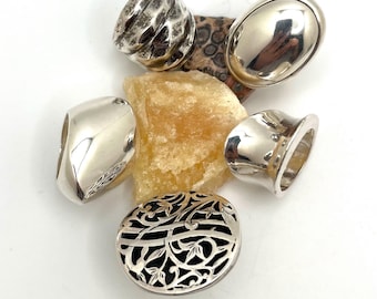 May include: An assortment of silver rings in various designs. One ring has a textured surface, another is oval-shaped, and a third features a floral filigree pattern. The rings are displayed on a white surface with natural stones.