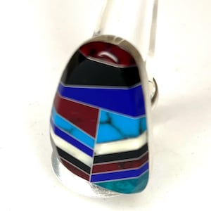 Puede incluir: Un anillo de plata con una piedra grande y colorida. La piedra presenta un diseño geométrico con secciones rojas, negras, azules, turquesas y blancas. El anillo está sobre un fondo blanco.