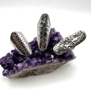 May include: Three silver-toned rings with distinct textures. The left ring features a repeating scale pattern, the center ring displays an engraved floral vine motif, and the right ring showcases intricate filigree work with floral elements.