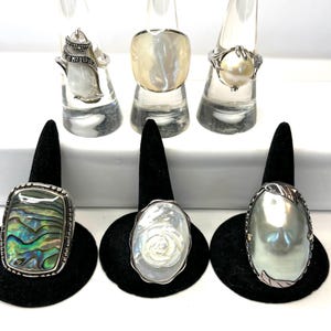 Puede incluir: Seis anillos en exhibición. Tres anillos están sobre soportes transparentes y tres sobre soportes negros. Los anillos presentan varios diseños, incluyendo una perla, abulón y nácar. Los anillos son de color plateado.