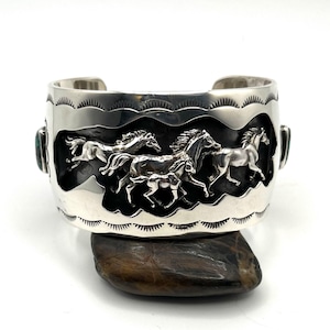 May include: A wide silver cuff bracelet with a detailed relief of running horses. The bracelet has a dark background and is accented with turquoise stones. The edges are decorated with a repeating pattern. This equestrian-themed jewellery piece is displayed on a dark stone.