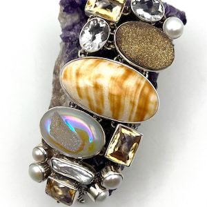 May include: A multi-stone bracelet featuring a large oval striped beige stone, a shimmering iridescent druzy cabochon, faceted yellow citrine, clear quartz, and white pearls set in silver-toned metal links, resting on a jagged purple amethyst cluster.