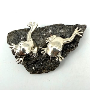 Peut inclure: Deux figurines de grenouilles en argent poli avec des surfaces lisses et réfléchissantes et des pattes palmées détaillées. Chaque grenouille est représentée en position rampante avec les membres étendus, affichant un éclat métallique.