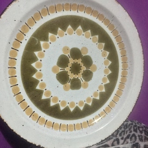 May include: A white ceramic plate with a decorative design. The plate features a central olive green circle with a floral pattern, surrounded by tan and olive green geometric shapes. The rim has a speckled texture.