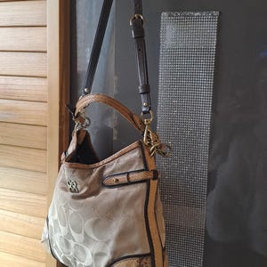 Könnte beinhalten: Beige Coach Handtasche mit sich wiederholendem Logo-Muster. Die Tasche hat braune Lederbesätze, einen Boden mit Schlangenmuster und einen braunen Schulterriemen. Goldfarbene Hardware und ein grünes Band ergänzen das Design.