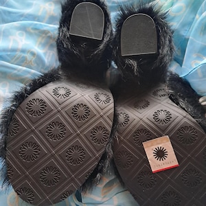 May include: Pair of black fur boots with a textured sole featuring a repeating floral pattern. The boots have a label attached with the brand name. The boots are on a blue patterned fabric.