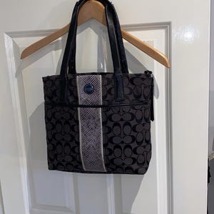 Puede incluir: Un bolso tote negro y gris con un patrón de logotipo repetido, asas de cuero negro y una franja vertical central con un estampado texturizado similar a la piel de serpiente. Un pequeño emblema circular azul está centrado en la parte superior.