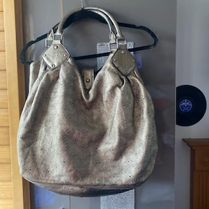 Puede incluir: Un bolso hobo de color dorado metálico con un patrón floral perforado en toda la superficie. El bolso cuenta con dos asas redondeadas unidas con herrajes plateados, una silueta fruncida y una lengüeta de cierre a presión en la parte superior.