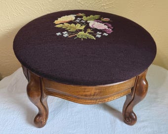 Taburete redondo de estilo vintage con cojín bordado con motivos florales y patas de madera curvadas.
