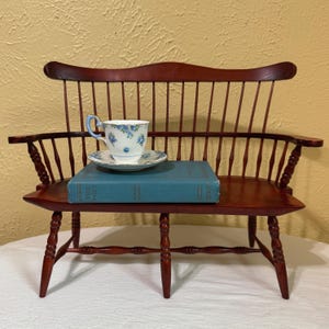 May include: A miniature wooden Windsor-style bench with a dark brown finish, featuring a curved backrest and spindle supports. A teal hardcover book titled The Poet sits on the seat, topped with a white porcelain teacup and saucer.