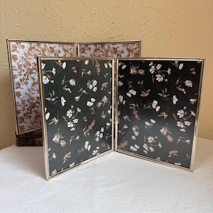 May include: A three-panel folding picture frame with a thin gold metal border. The two front panels feature a dark green background with a white and light pink floral pattern, while the back panel displays a light cream background with floral prints.