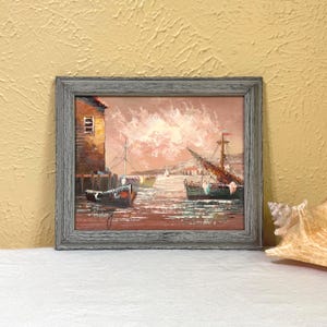 May include: A framed oil painting of a harbor scene with boats and a building. The painting features a muted color palette of browns, reds, and blues. The frame is gray.