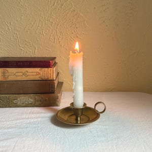 Può includere: Una candela bianca accesa in un portacandele in ottone con base rotonda e manico. La cera della candela si sta sciogliendo e la fiamma è luminosa. Una pila di libri antichi è sullo sfondo.