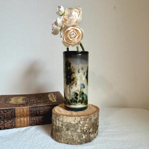 Puede incluir: Un jarrón cilíndrico de cerámica con un diseño de paisaje, con árboles, pájaros y flores. El jarrón se exhibe sobre una rodaja de madera y contiene varias flores de color claro. Dos libros antiguos son visibles en el fondo.