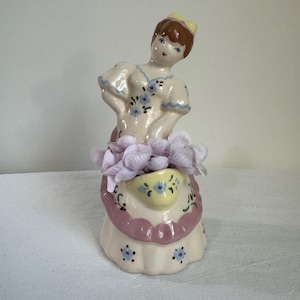Puede incluir: Figura de cerámica vintage de una mujer con un vestido, sosteniendo una cesta de flores. La figura está pintada con colores pastel, incluyendo rosa, azul y amarillo. La mujer tiene cabello castaño y una corona amarilla. La cesta está llena de flores moradas claras.