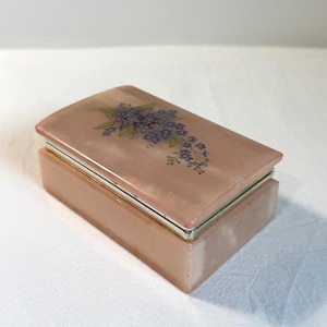May include: A rectangular, pink jewelry box with a hinged lid. The lid is decorated with a floral design of blue flowers and green leaves. The box has silver-colored metal trim.