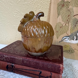 May include: A brown ceramic acorn-shaped container with a textured surface and a stem. The container sits atop two antique books with burgundy covers. The background includes a painting of a bird and leaves.