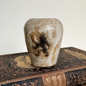 May include: A small, handcrafted ceramic vase with a textured, off-white and brown glaze. The vase features a dark brown octopus design on the front. The vase is sitting on top of a vintage book.