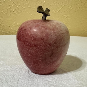 May include: A polished, rounded apple-shaped sculpture crafted from mottled pink stone. The object features a smooth, glossy surface with natural variations in color and a dark, metallic-toned stem attached to the top center.