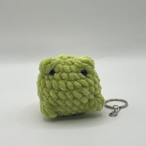 Op de afbeelding: Een handgemaakte, lichtgroene gehaakte kikker sleutelhanger. De kikker heeft een kubusvorm met zwarte ogen. De sleutelhanger heeft een zilverkleurige metalen ring en sluiting. De kikker is gemaakt van zacht, getextureerd garen.