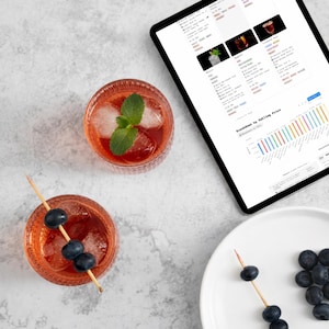 Peut inclure: Vue aérienne de deux cocktails dans des verres ronds avec de la glace et des feuilles de menthe. Un verre contient des myrtilles sur une brochette. Une tablette affiche une page web avec des données. Une assiette avec des myrtilles et le texte "Cocktail Lab" sont également visibles.