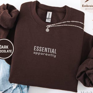 May include: A dark chocolate brown crewneck sweatshirt featuring the embroidered text ESSENTIAL apparently in a light grey font. The garment has long sleeves and a ribbed collar, displayed alongside blue denim jeans and a silver chain necklace.