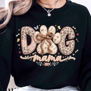 May include: Black sweatshirt with the words "DOG mama" in a decorative design. The letters "D" and "G" have a leopard print pattern, and the paw print is decorated with a bow and mushrooms.